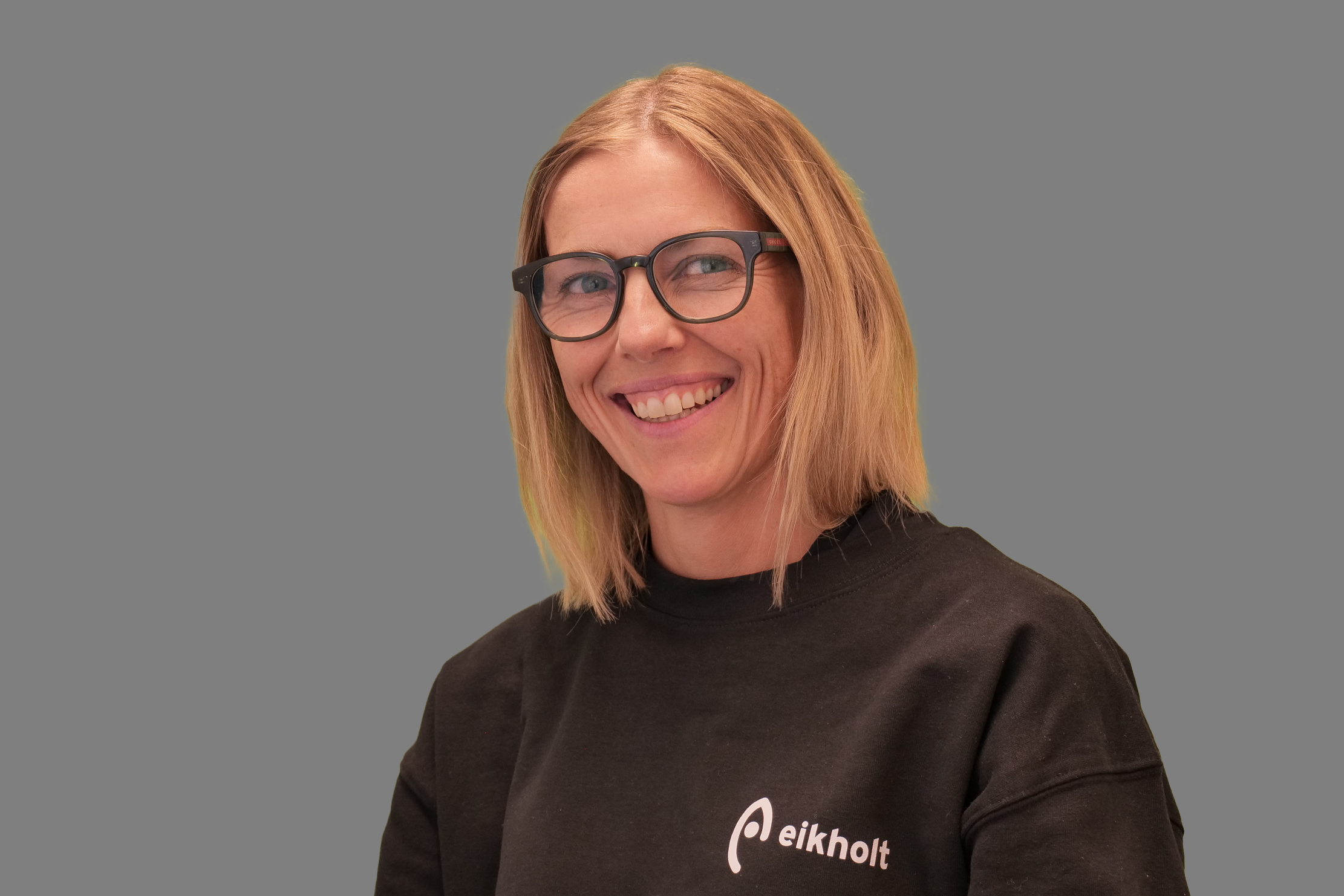 Tanja has light-coloured hair down to her shoulders. She wears glasses and smiles with her mouth open. She is wearing a black jumper with the text "Eikholt"