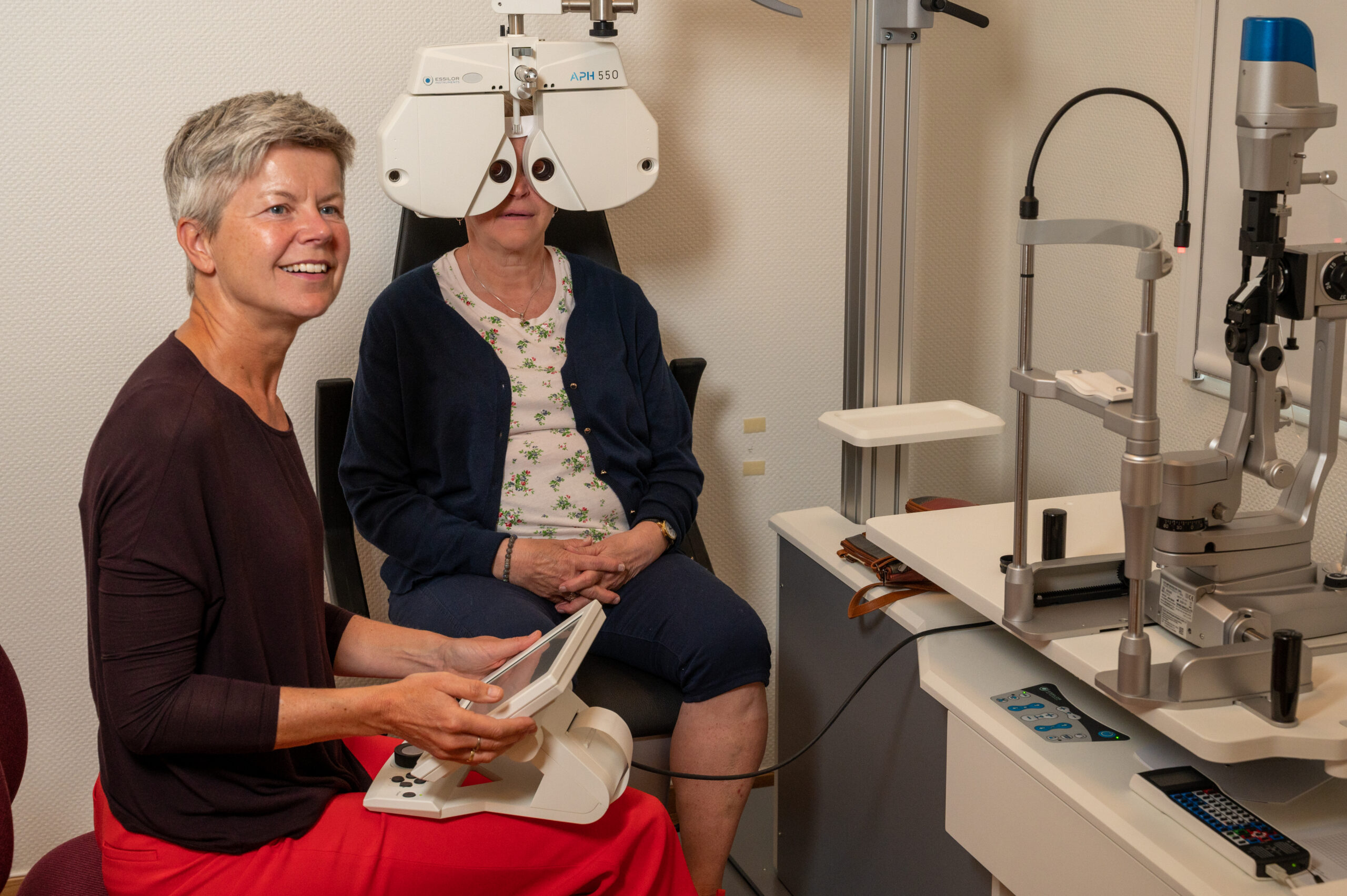 To personer sitter inne i AV-klinikken,. En holder et apparat på fanget. En ser inn i et Phroropter.