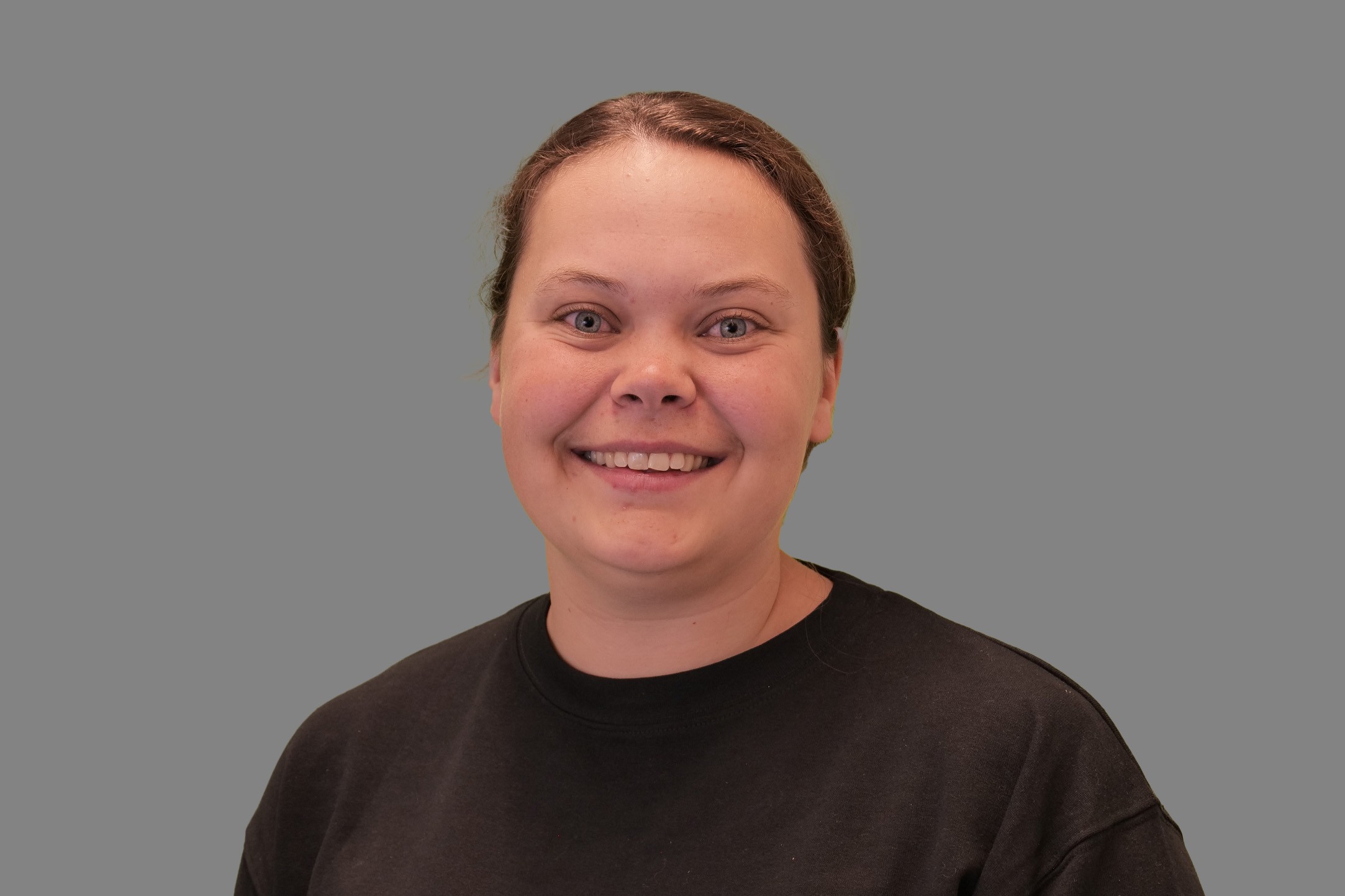 Anne-Mayn muotokuva. Hänellä on tummat hiukset, jotka on sidottu poninhännäksi. Hänellä on musta pusero ja hän hymyilee.