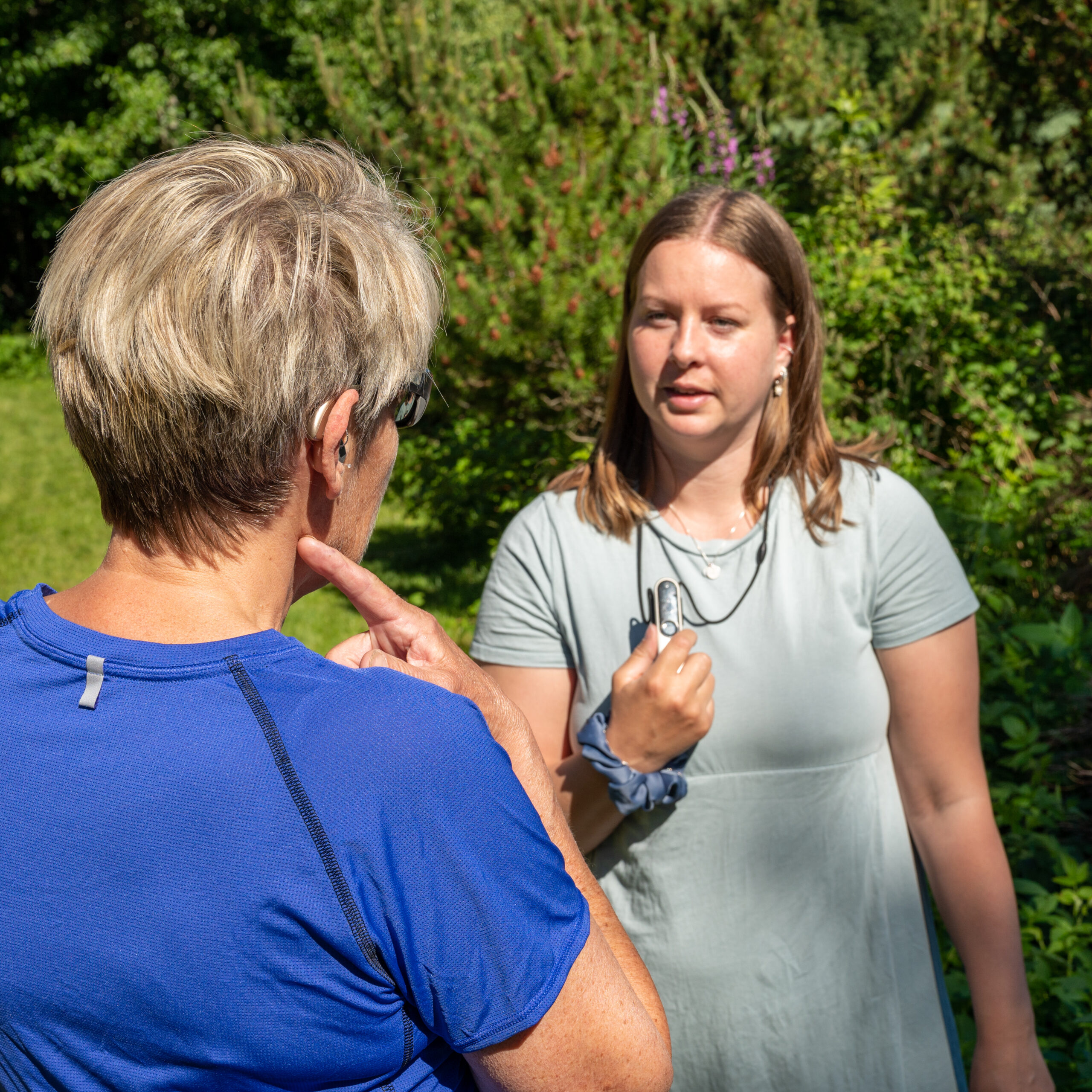 To personer står utendørs mot hverandre. Den ene står med ryggen til, har grått hår, t-skjorte, høreapparat og filterbriller. Den andre personen har blondt hår til skudrene, står med en roger-penn rundt halsen og har på kjole. Rund vises grønn natur.