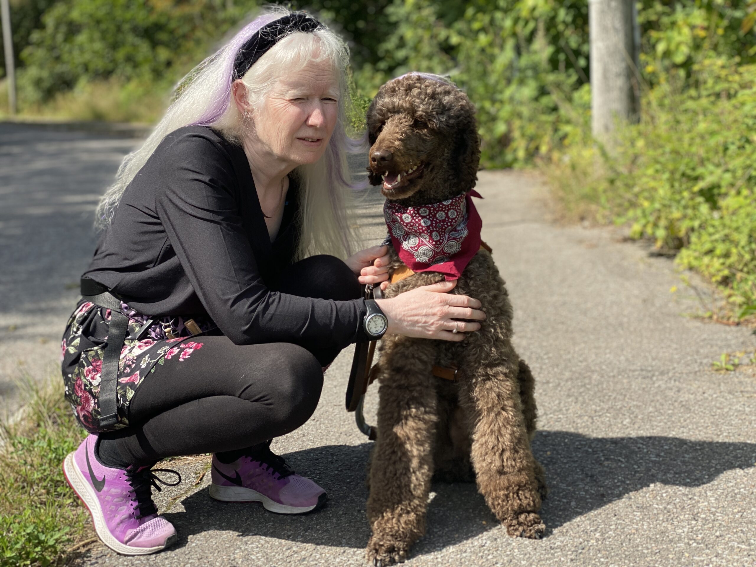 En person sitter på huk ved siden av førerhunden sin på en vei. Rund de er det trær. Hun har langt lyst hår, hårbånd, sorte klær og joggesko. Hunden har skjerf, er brun og krøllete.
