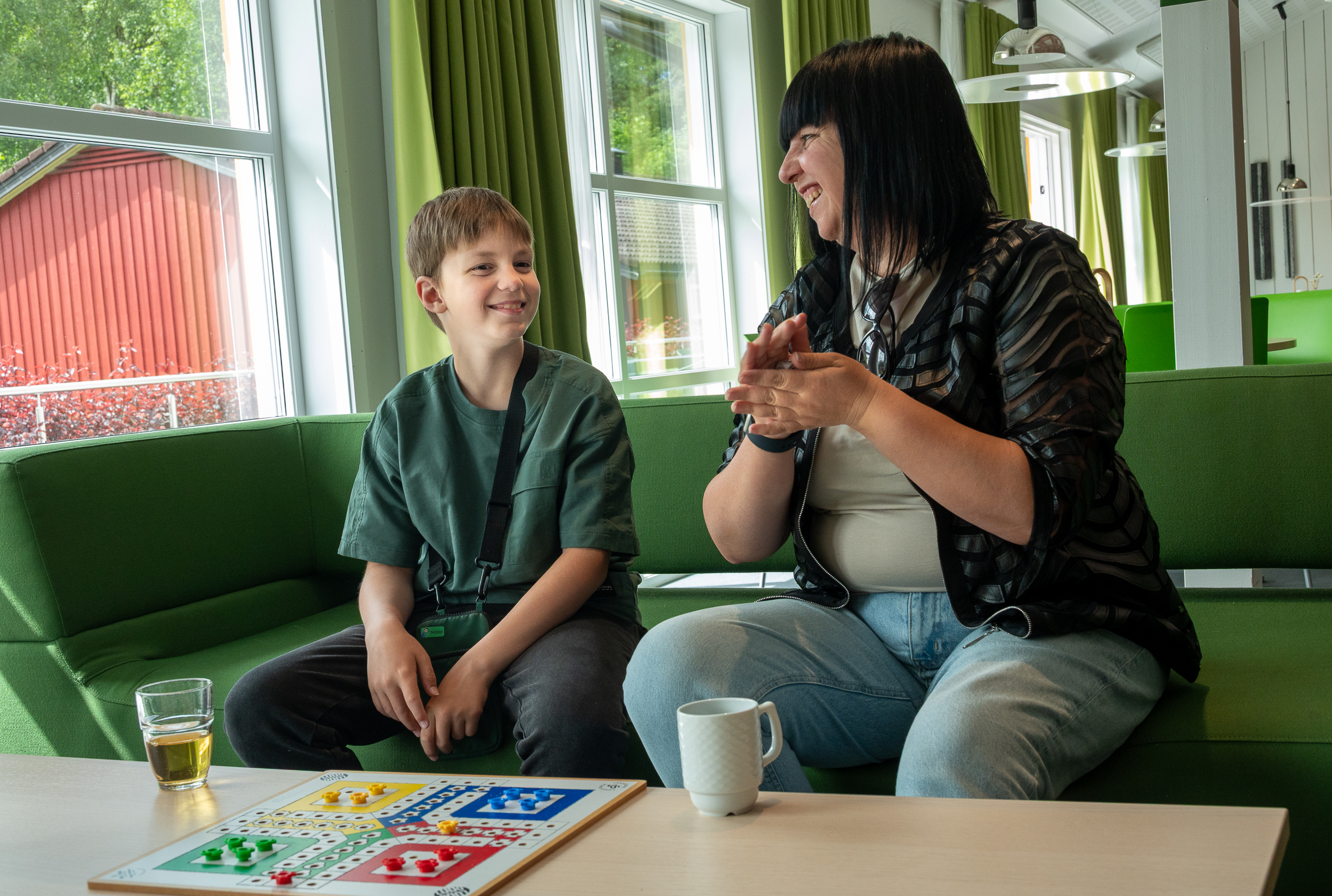 En gutt og en dame sitter ved et bord inne og spiller ludo. De smiler mot hverandre.