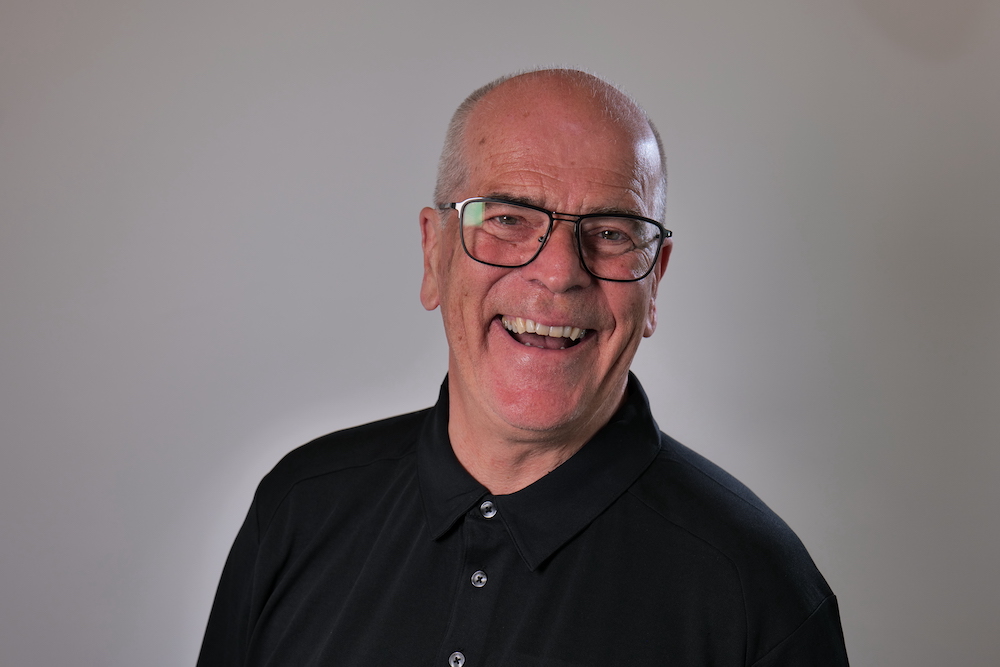 Rolf har kort grått hår. Han bruker mørke briller og sort skjorte med knapper. Han har et stort smil. 