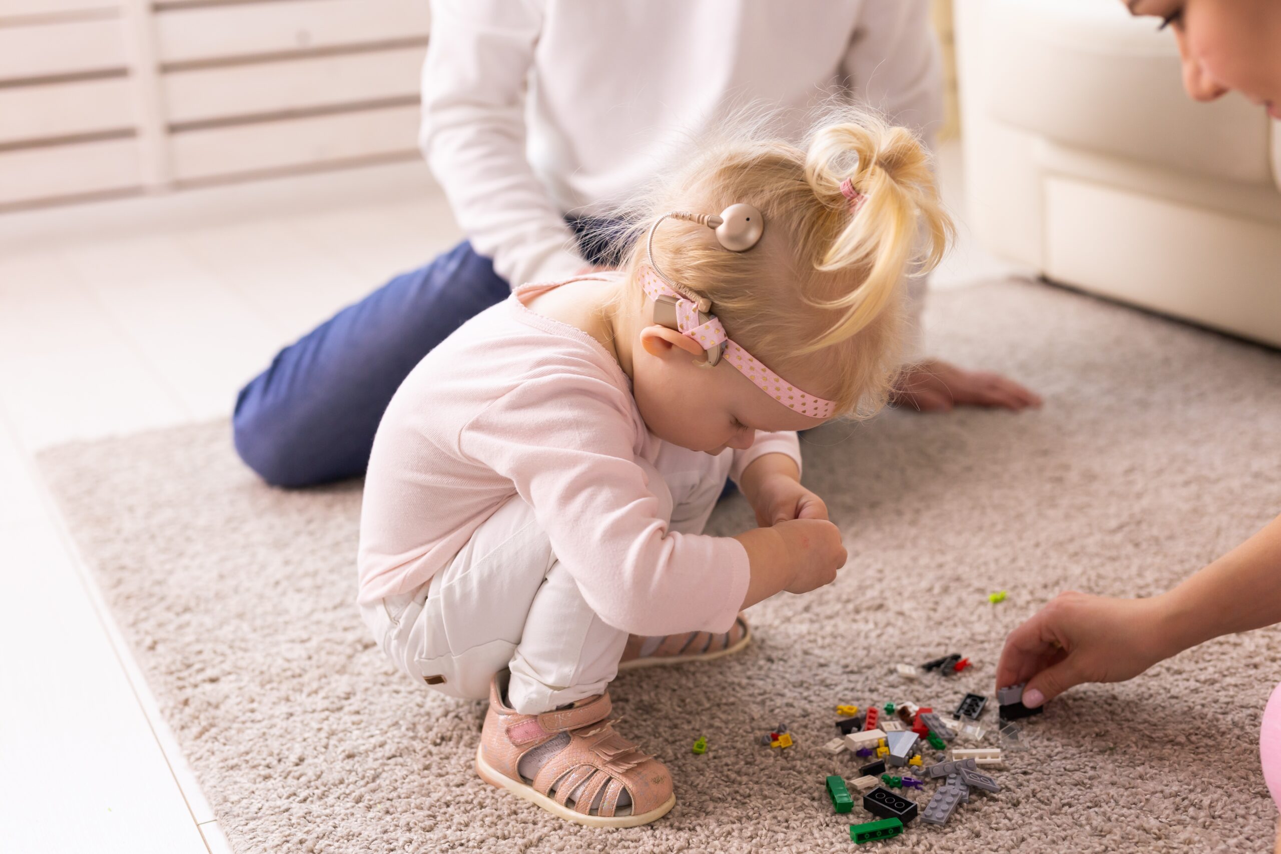 En liten jente sitter på huk på gulvet. Hun er kledd i rosa og hvitt, har hårbånd og cochlea implantat. Hun leker med lego med to andre personer .