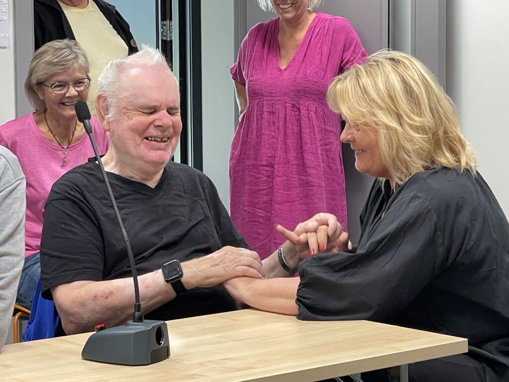 Harald Vik sits smiling at a table. He is holding an interpreter in his hands. In front of him is a microphone, in the background are people.