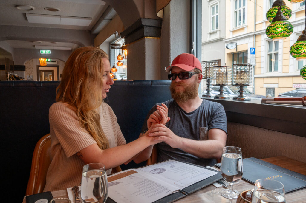 Två personer på en restaurang. Den ena har keps och filtglasögon. De samtalar med taktil kommunikation.