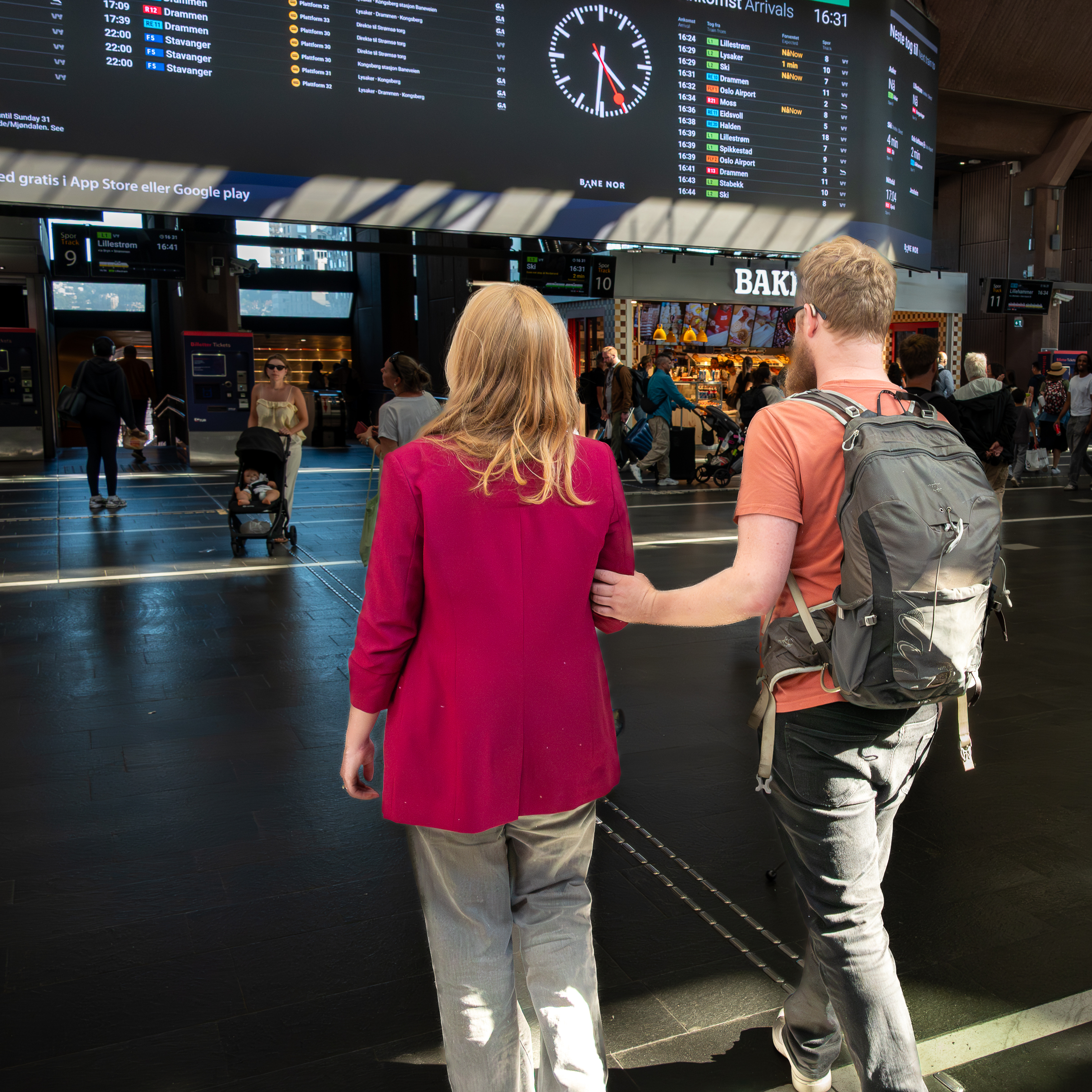 To personer går på Oslo, Sentralstasjonen i avgangshallen. Se ser mot en opplysningstavle. Den ene holder i albue, har briller, høreapparat og sekk på.