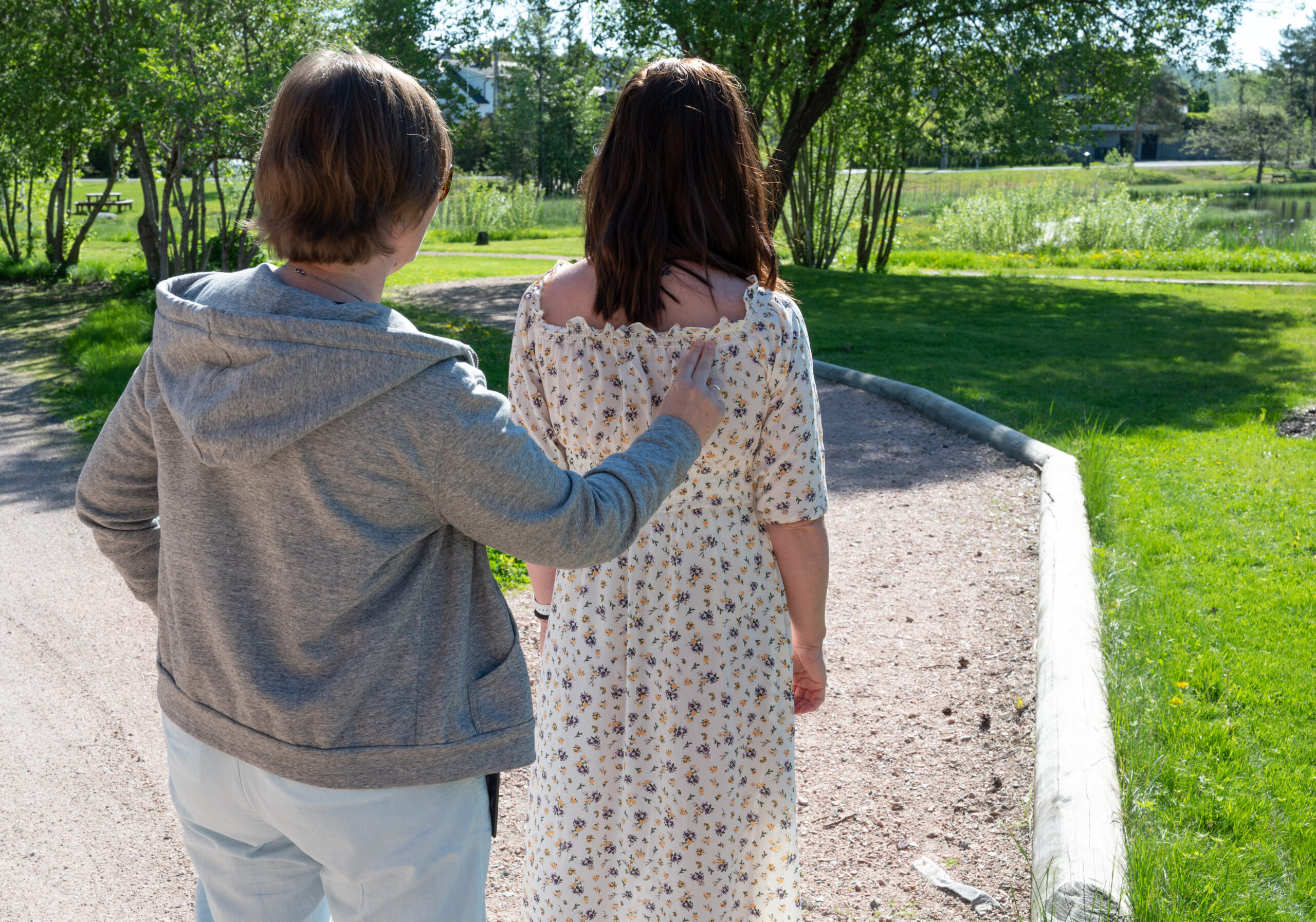 To personer står ute. En person holder to fingre på ryggen til den andre. De ser ned mot en sti. Ved siden av er det grønt gress.