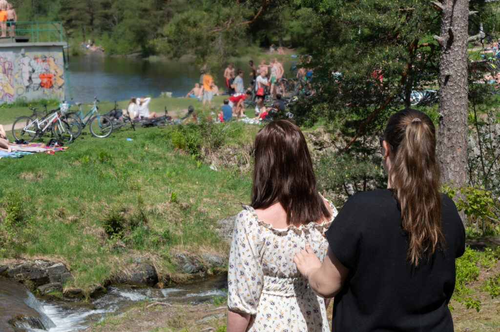 To personer står og ser utover et vann. I vannkanten er det mange mennesker.  En person gir en annen haptiske beskrivelser av det de ser .