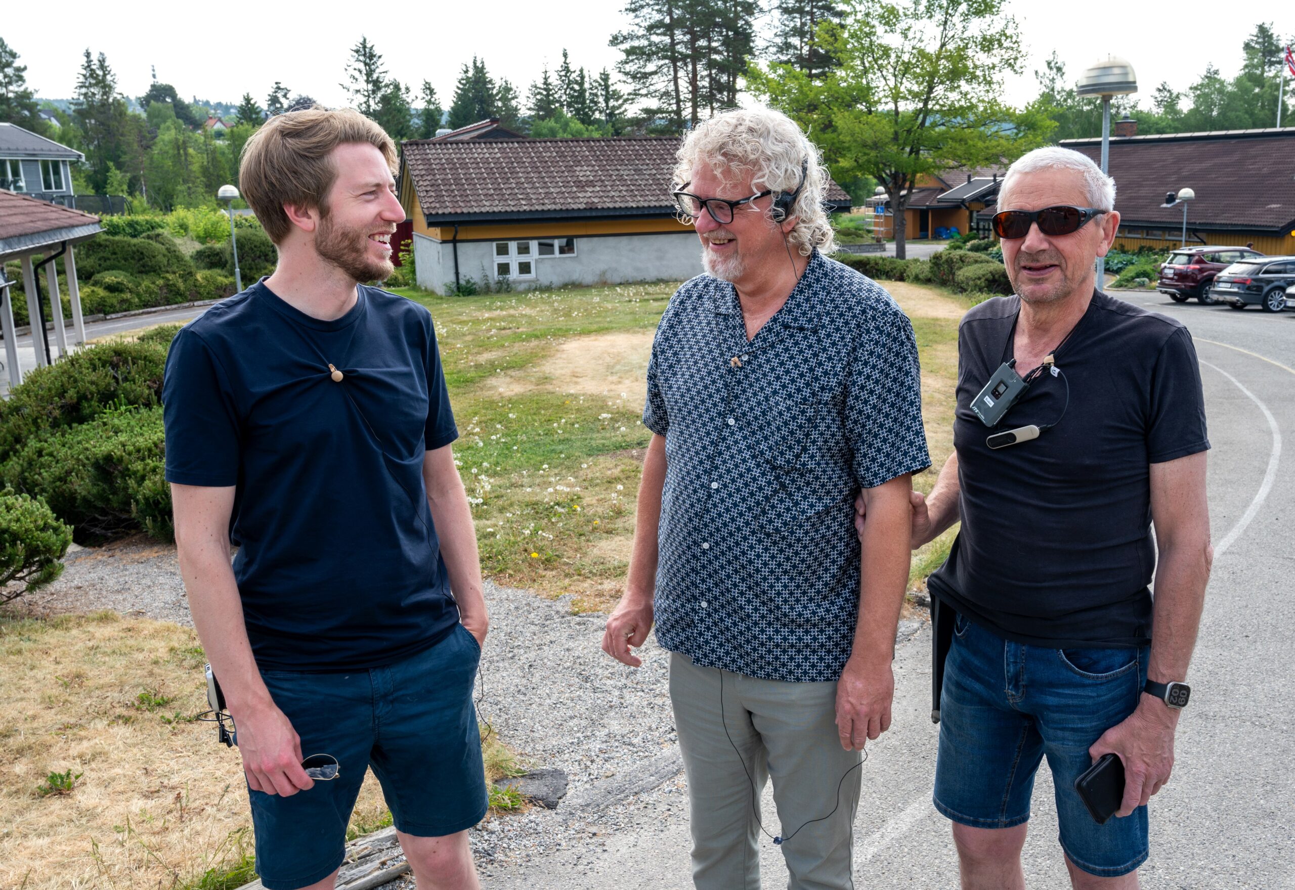 Tre personer står utendørs. En har på seg briller, rundt halsen henger hørselsteknisk utstyr. En annen har på seg headsett. De smiler. Bak vises hus, gress og asfalt.