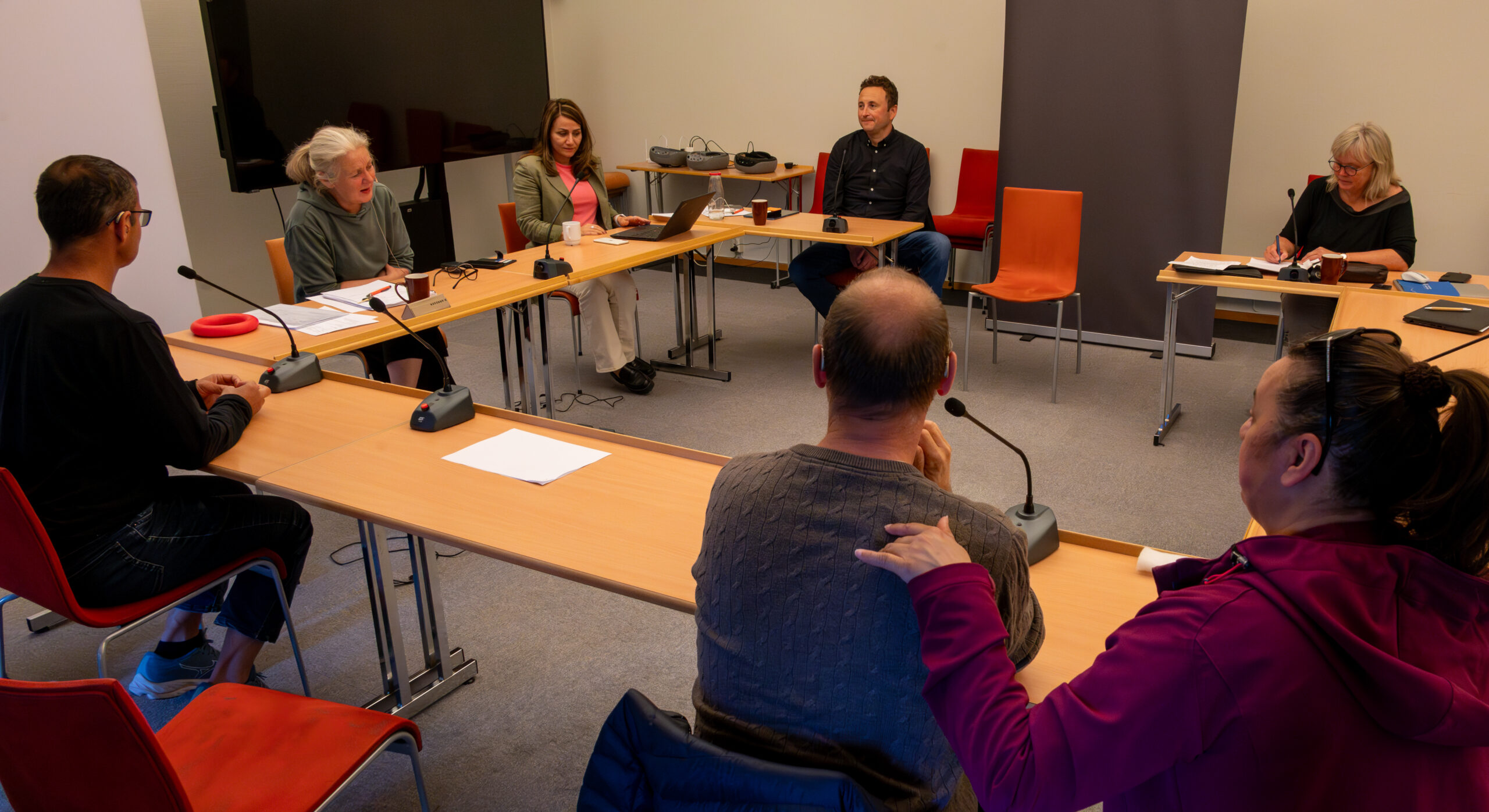 Flere personer sitter samlet rundt et bord og diskuterer. Det er mikrofoner på bordene.