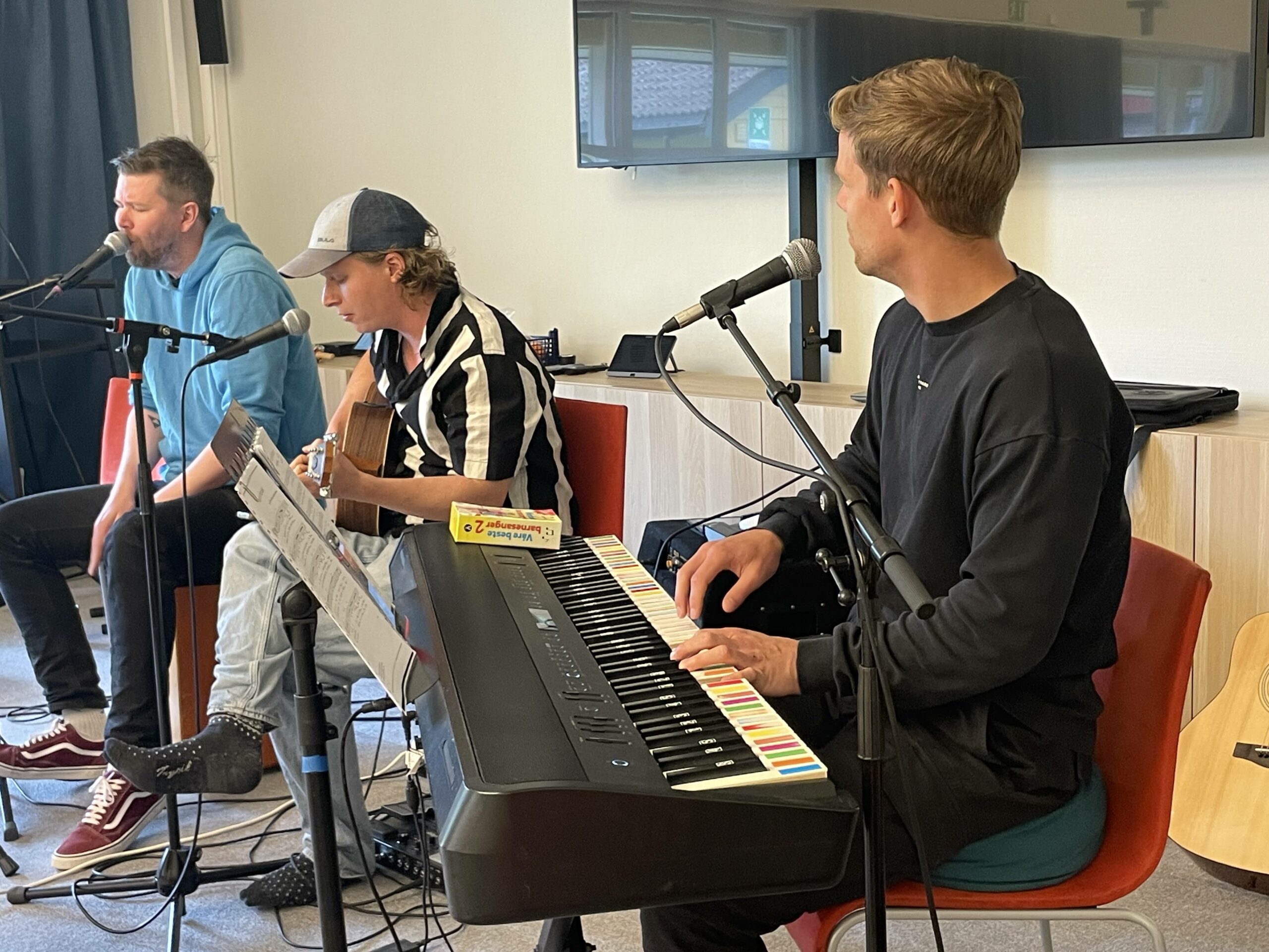 Tre musikere spiller gitar og keyboard og synger i mikrofoner innendørs