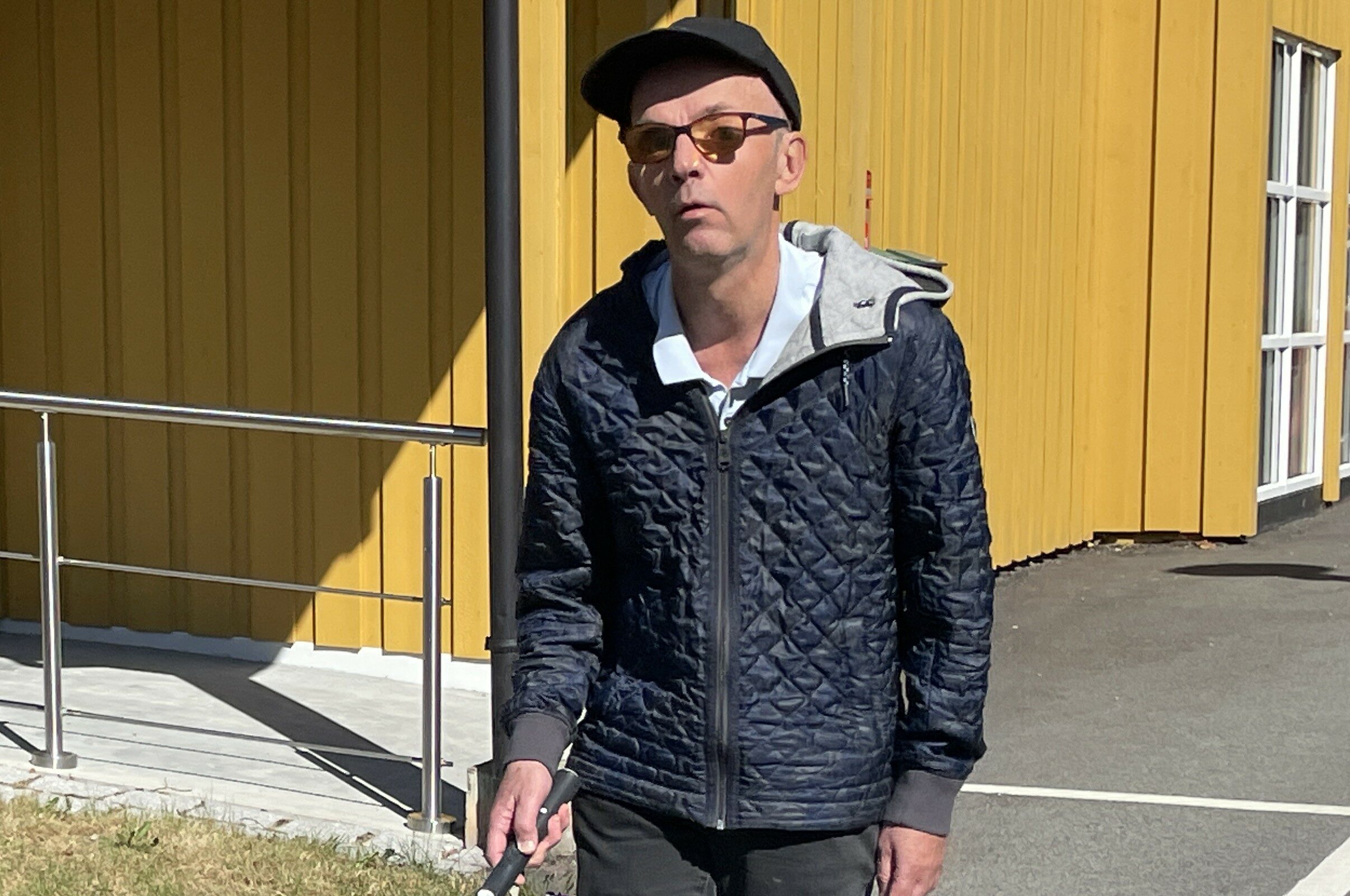 Mann med solbriller og mobilitetsstokk utenfor en gul bygning