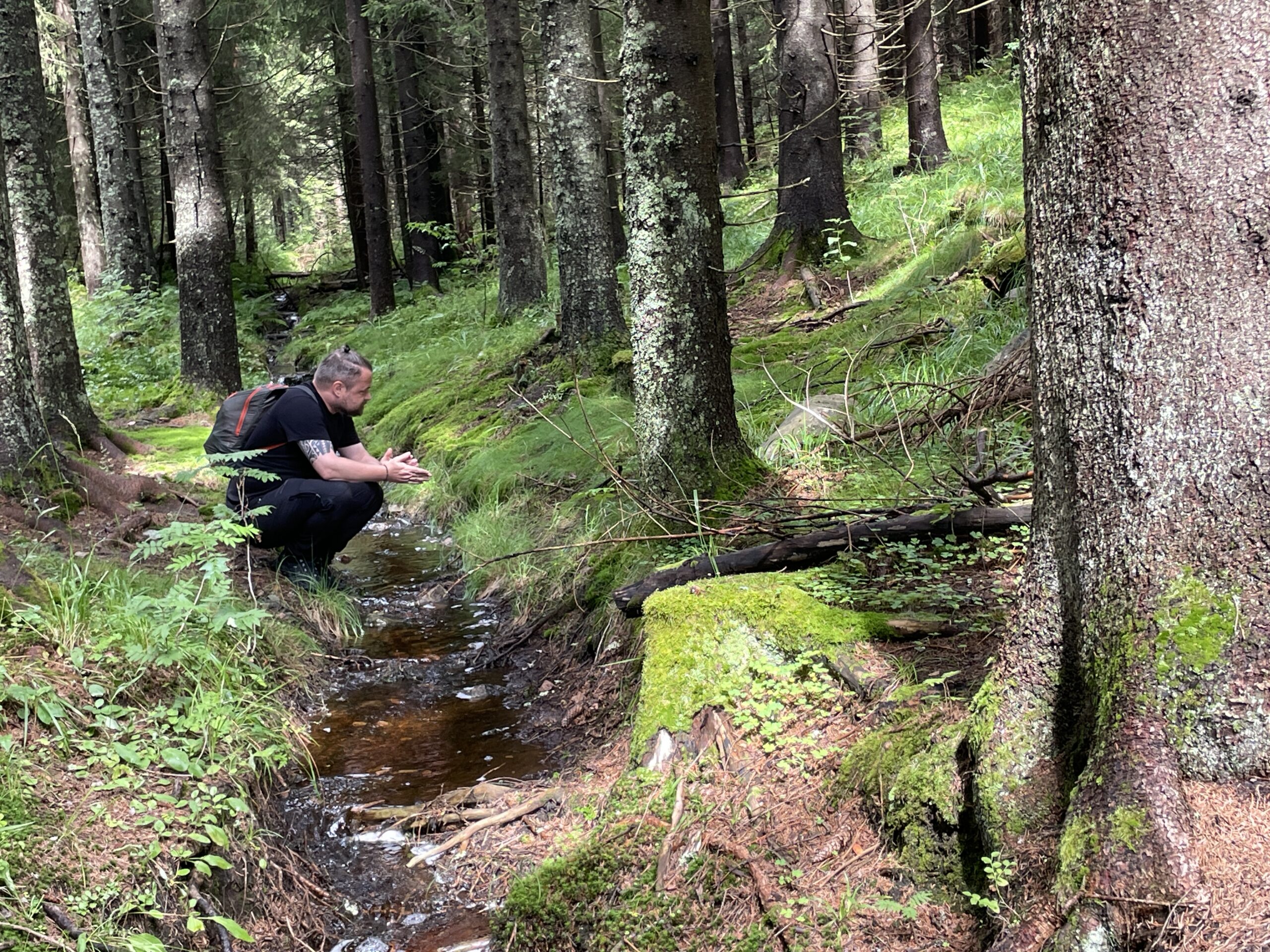 En person sitter på huk ved en bekk i skogen. Rundt er de trær, mose og solskinn. Personen har sekk og ser tankefullt foran seg.