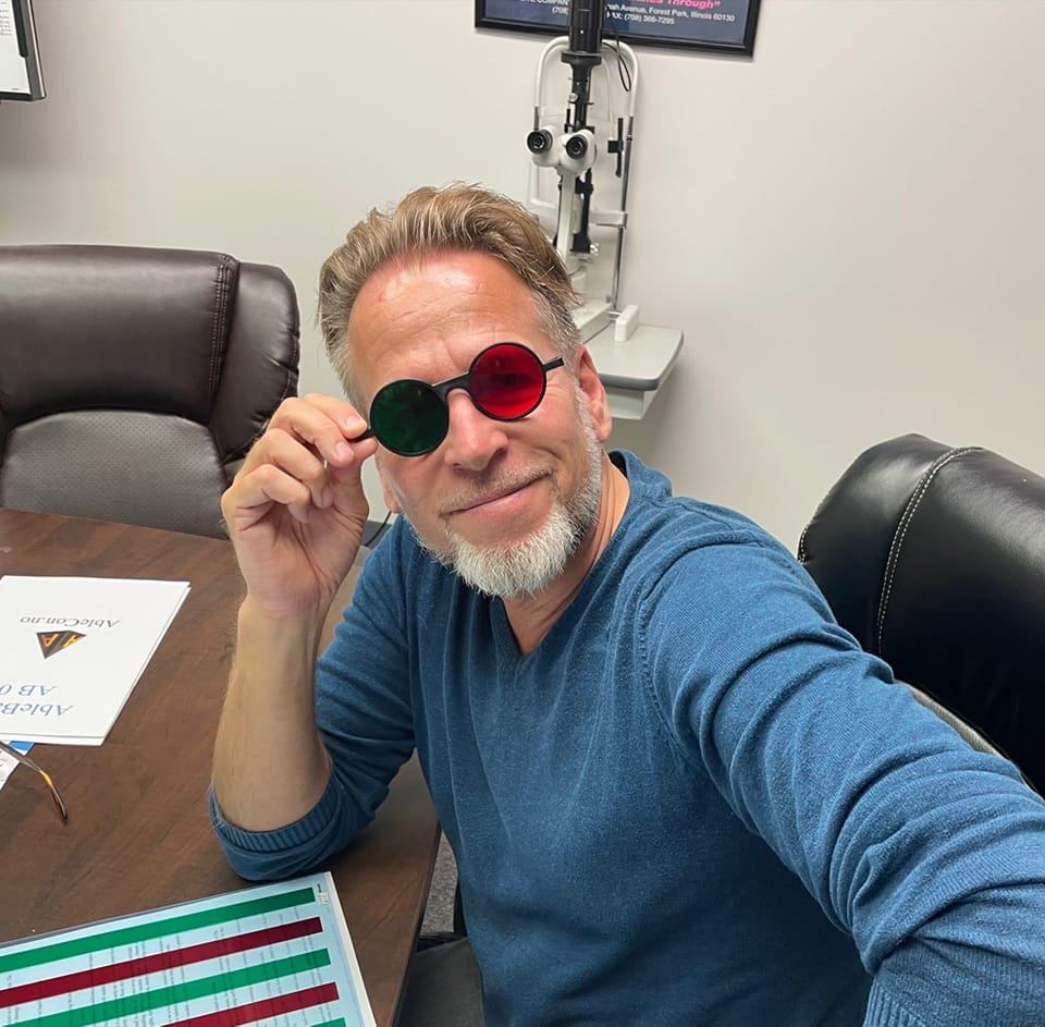 Picture of Egil Edwin. He has short blonde hair, a grey beard. He is sitting in a chair, wearing a pair of glasses with different coloured lenses. He is holding his hand on one of the temple arms of the glasses with his elbow on the table in front of him.
