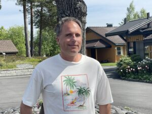 Marius Langeland Myklebust is standing outside. He has short hair, a sly smile and is wearing a white t-shirt. Buildings and trees in the background.
