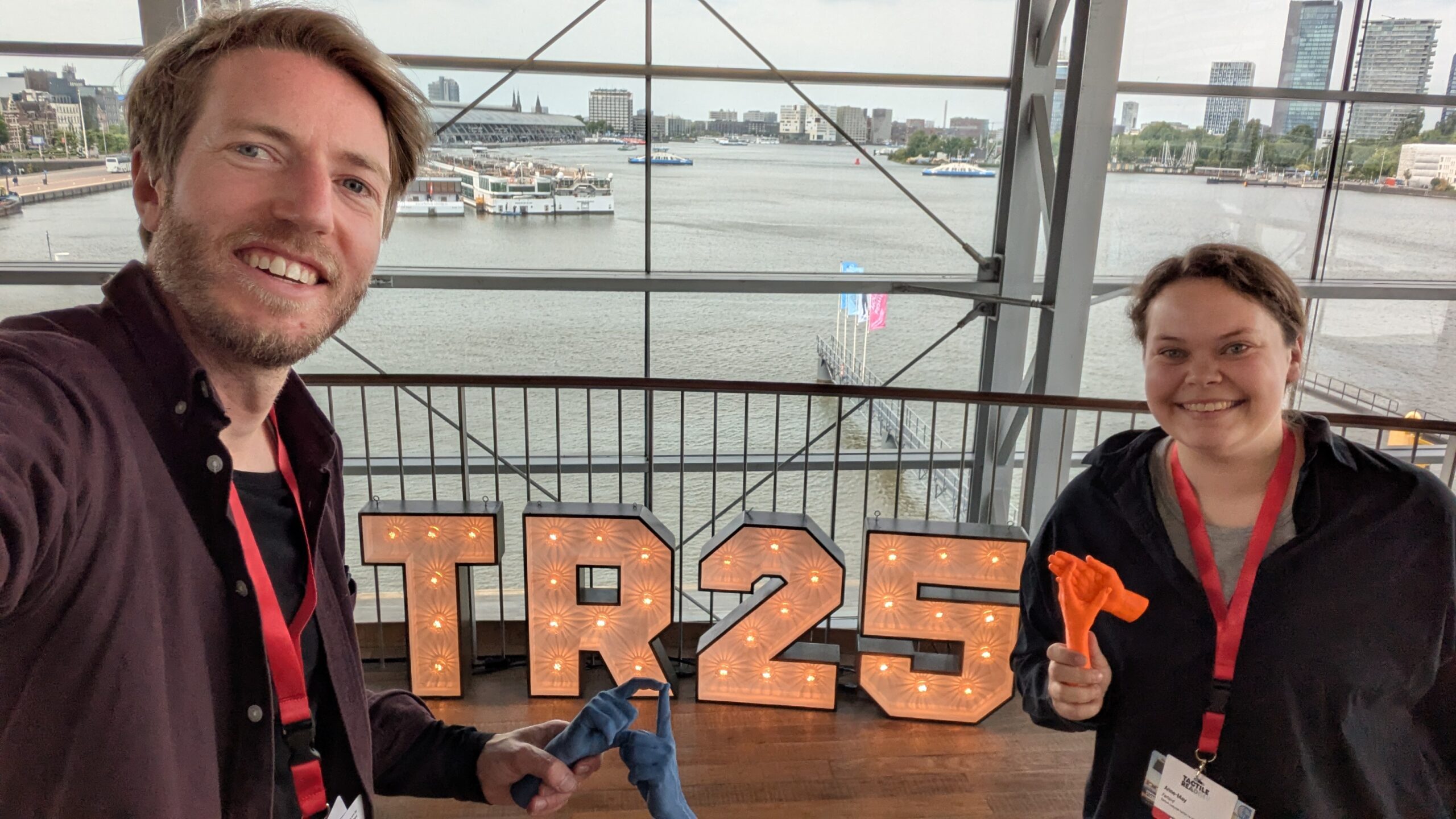 Rolf Mjønes og Anne May førland sitter ved et bord med 3D-printede hender. På bordet står også store bokstaver med TR25 som står for Tactile Reading 2025. I bakgrunnen vises en kanal med båter.