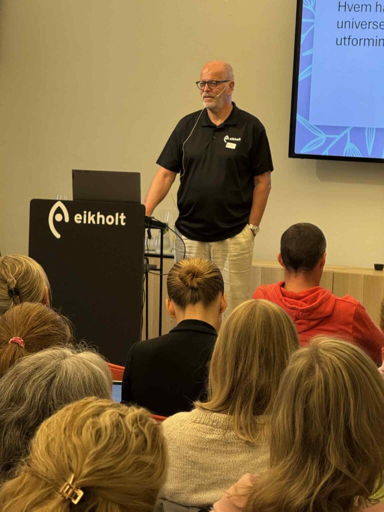 Arne Kjeldstad står ved en talerstol. Han holder en hånd på en PC. Foran han sitter mange mennesker.