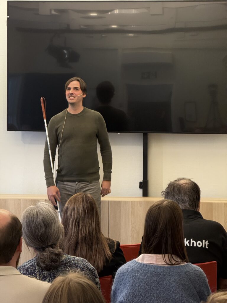 Thomas Tvedt står med en mobilitetsstokk i ene hånden. Foran han sitter mennesker, bak han henge en TV-skjerm.