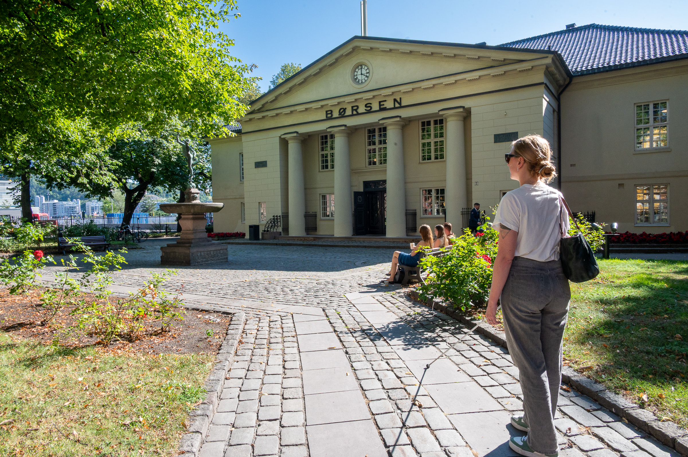 En person står utenfor Oslo børs. Personen har skulderveske, mobilitetsstokk, briler og høreapparat. Foran sitter tre personer på en benk.