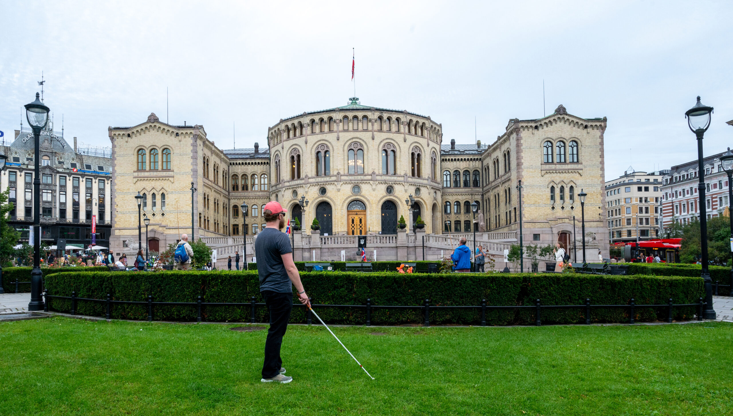 En person står utenfor stortinget. Personen har caps og mobilitetsstokk. Gress og hekk vises foran bygningen.
