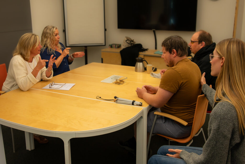 Fire personer ved et bord, to og to sitter mot hverandre. To stk bruker tegn og de to andre ser mot disse. bak den ene sitter også en person og gjjør tegn på ryggen. Det ligger en mobilitetsstokk på bordet.