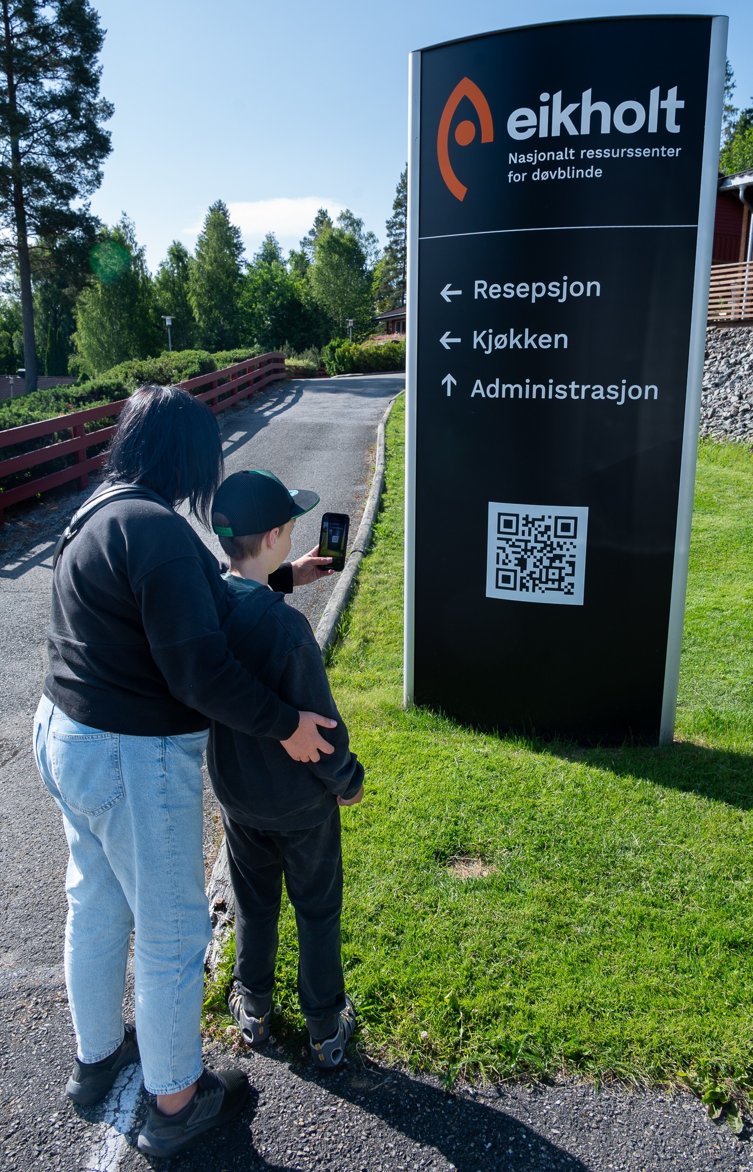 En gutt holder en mobil mot en QR-kode på et skilt. Bak står moren og holder rundt han.