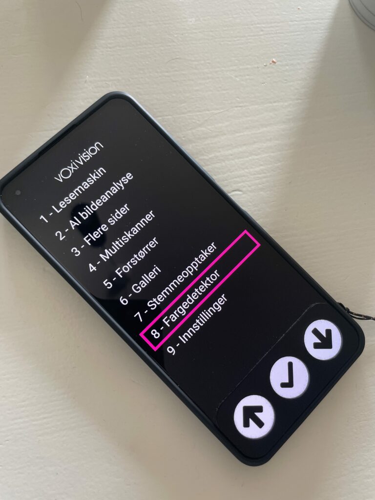 VoxiVision, et hjælpemiddel på størrelse med en smartphone. Skærmen viser tekst, og nedenunder er der tre store runde navigationsknapper med symboler for op, ok og ned.