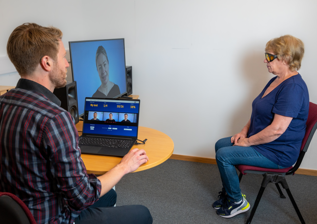 en mann sitter foran en laptop, i bakgrunnen en dame som ser på en TV-skjerm