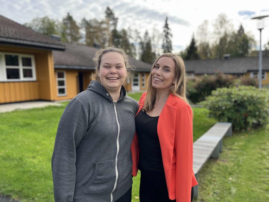 Anne-May and Victoria are standing outside. Anne-May is smiling and wearing a grey jacket, Victoria is smiling and wearing an orange jacket and a black sweater. In the background, houses and buildings.