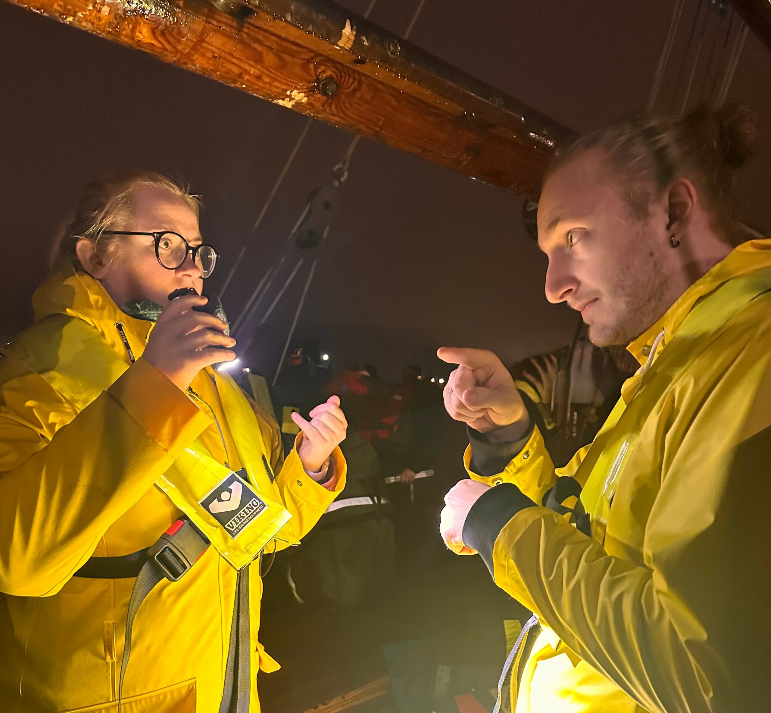 To mennesker står på en båd. Det er mørkt. De har gult regntøj på. Den ene lyser med en lommelygte på den anden, som bruger tegnsprog.