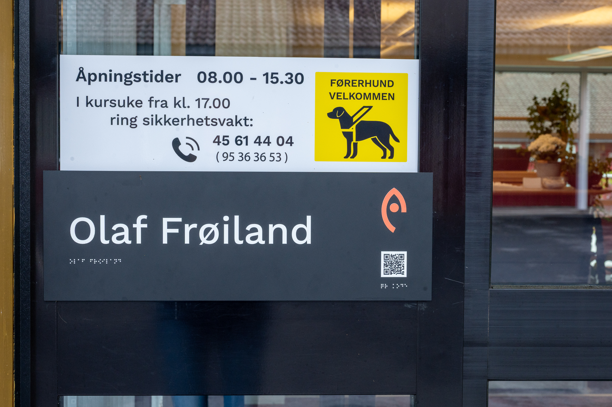 Skilt med åpningstid: 08:00 - 16:00, førerhund velkommen og navnet på bygget: Olaf Frøilands hus i sortskrift og punkt. Det er ogsåen Qr-kode.