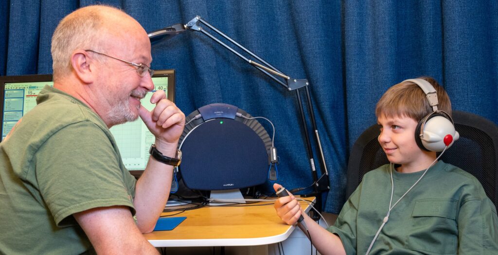 En voksen og et barn sitter og ser på hverandre og smiler. Barnet har på hodetelefoner. De sitter ved et bord med PC og hørselsapparater.