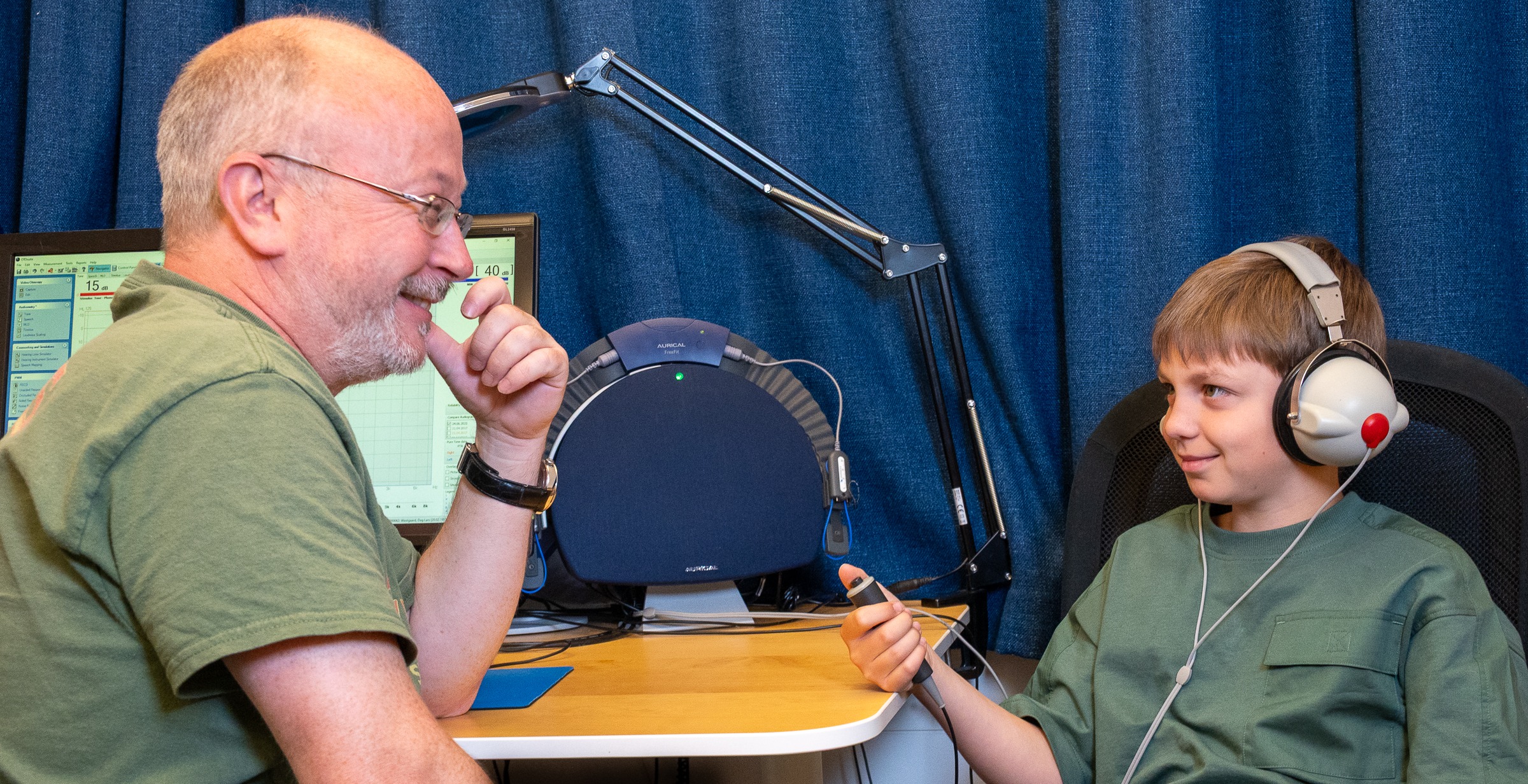 En voksen og et barn sitter og ser på hverandre og smiler. Barnet har på hodetelefoner. De sitter ved et bord med PC og hørselsapparater.