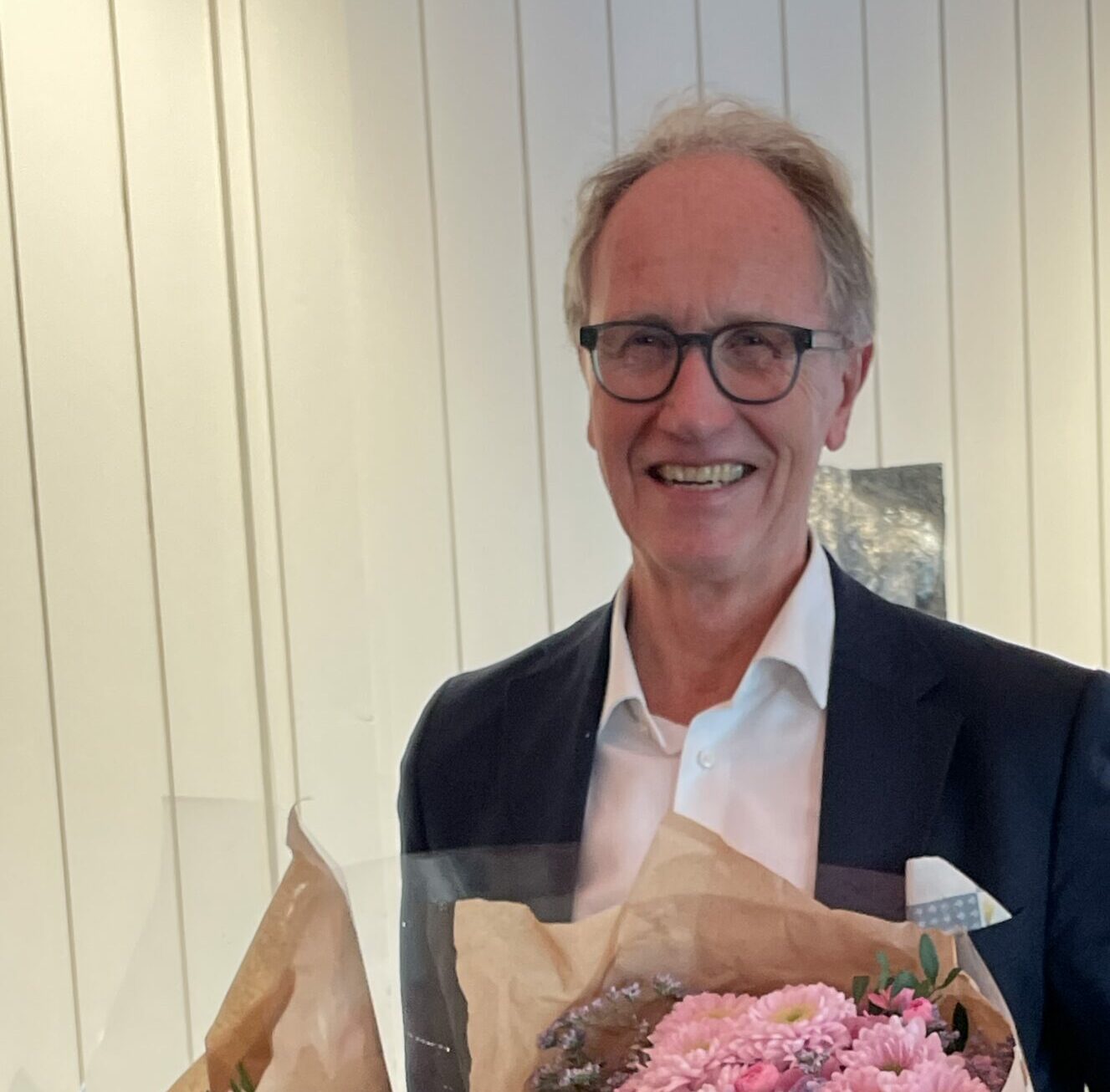 Roar Meland står innendørs med to blomsterbuketter i hånden. Han har dressjakke, skjorte og dongeribukse.