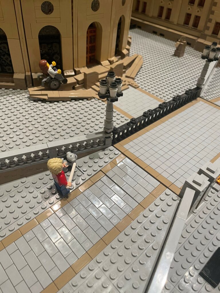 Legofigur med mobilitetsstokk og førerhund står utenfor porten på legobygningen av stortinget. En legofigur av en person i rullestol er på vei opp en rampe.