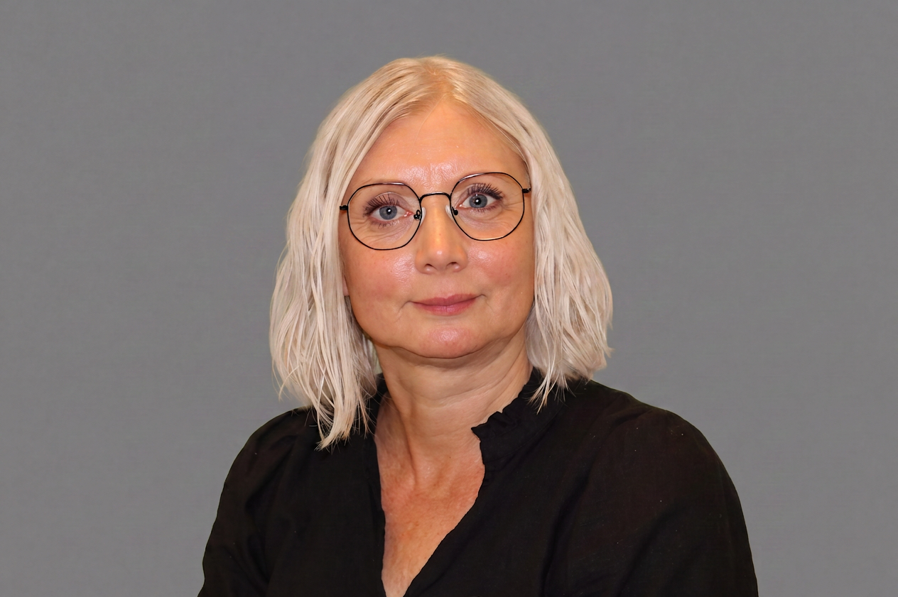 Jill har blondt hårtil skuldrene og briller. Hun bruker en sort skjorte.