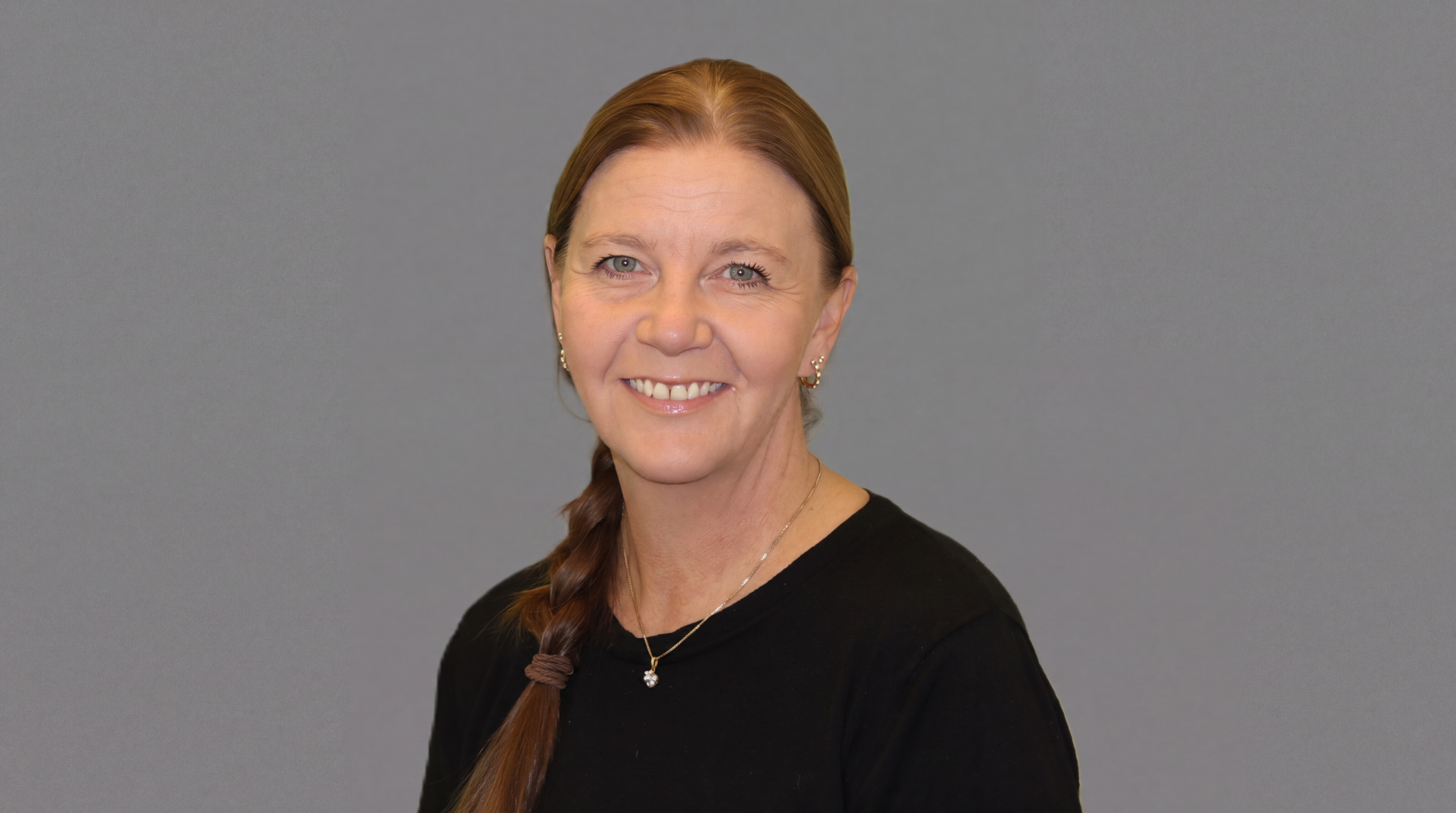 Portrait of Veronica Samuelsen. She is smiling at the camera, sitting slightly at an angle. Veronica has long brown hair braided into a plait that lies on one side. She is wearing a necklace and a black jumper.