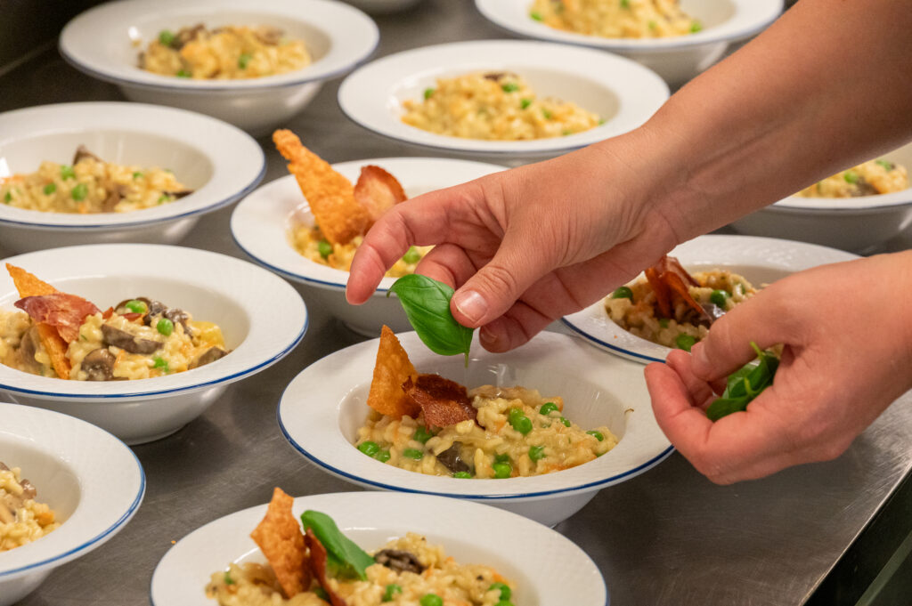 Skåler med mat på en benk. Det ser ut som risotto med erter, pyntet med basillikum og kjeks. Hender pynter maten med basillikum.