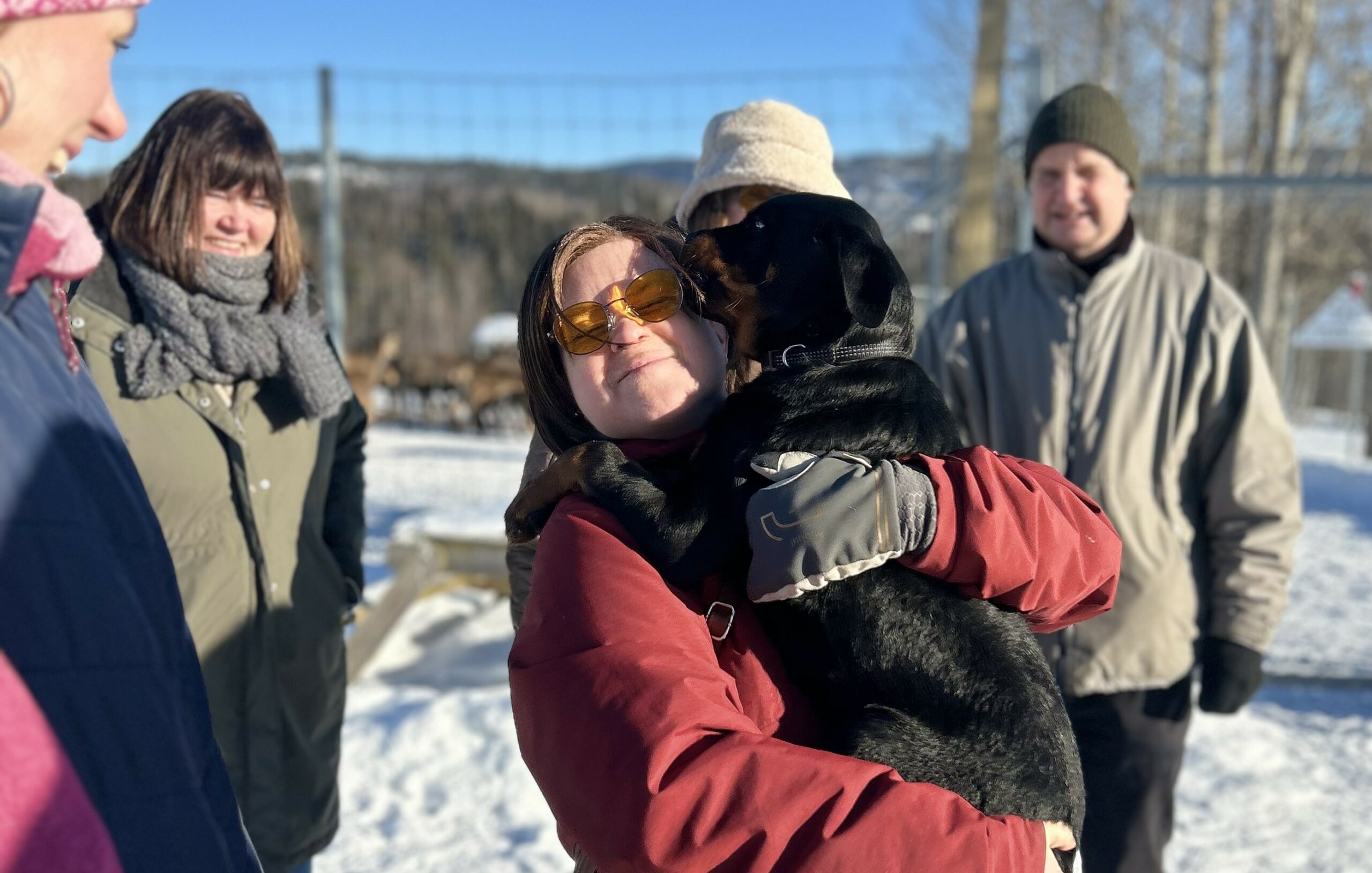 En person står ute med en heund i fanget. Flere personer står rundt. Det er vinter, snø og blå himmel. Hunden slikker jenta i ansiktet, hun lager grimase ved å knipe øyne og munn sammen.