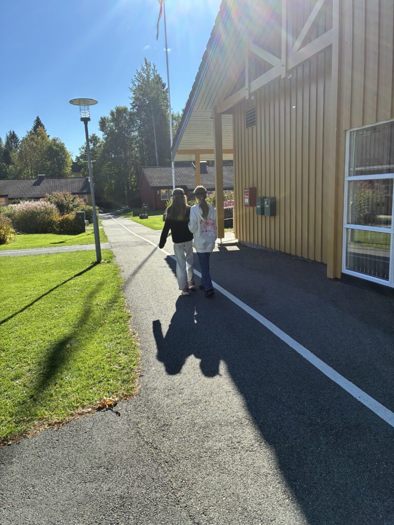 To børn går på en asfalteret gangsti langs en trebygning i solskin